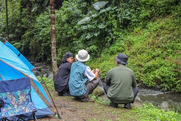 Fototapeta premium Three Friends Enjoying Peaceful Camping Trip In Lush Green Forest Near Tranquil Stream