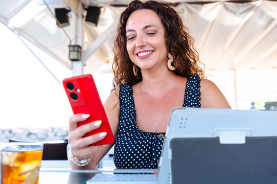 Smiling woman using smartphone with red case while working on tablet at outdoor cafe, modern lifestyle with digital technology, communication, leisure, business, internet connection, summer casual - Powered by Adobe