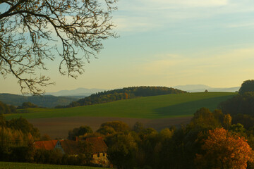 Peaceful autumn countryside scene with rustic farmhouse, vibrant fall foliage, green and brown farmland, rolling hills and distant mountains in soft golden evening light