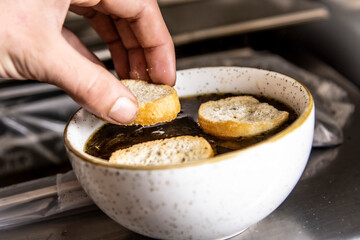 A close-up shot capturing a hand carefully adding toasted bread to a rich, warm onion soup. This image perfectly conveys the process of preparing a comforting and classic dish.