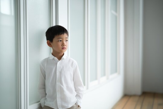 Thoughtful asian schoolboy in white shirt standing near window in bright hallway. Young male elementary student looking away indoors in quiet modern school