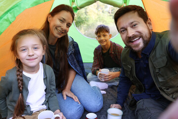 Happy family taking selfie in camping tent with mugs of hot drink outdoors