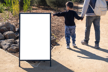 Blank white advertising mockup board on outdoor park pathway with an Asian child and adult parent walking hand in hand in the background. Concept of copy space, signage, family-friendly information.