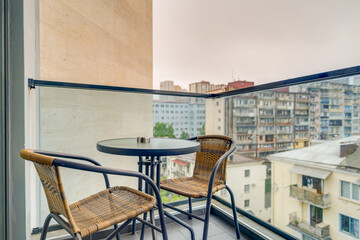 A clean balcony scene featuring wicker chairs and a small round table, with a glass railing and a distant urban skyline.