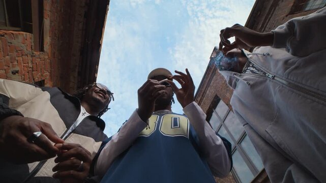 Low angle view of three young Black men standing in side street, smoking cigarettes while hanging out together