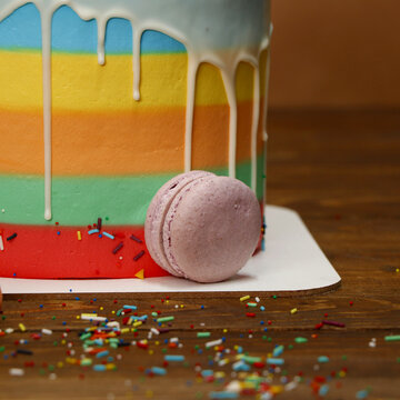 Rainbow layer cake decorated with colorful dripping icing and scattered sprinkles on wooden surface. A single pink macaron rests at the base of the cake adding contrast and elegance.