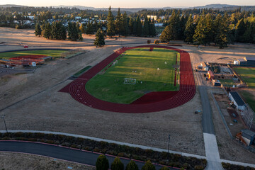 Aerial drone picture of college sports fields in Oregon City with soccer, baseball and track areas, students practicing on green lawns during fall sunset, surrounded by trees and campus buildings