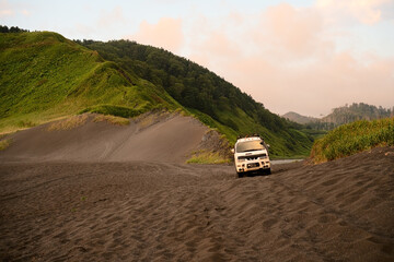 Fototapeta premium 4x4 adventure car parked on secluded sandy beach between green mountains and ocean waves. Perfect for overland travel and wilderness exploration concepts.