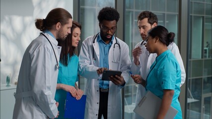 Multicultural five diverse people Caucasian Indian African women men doctors nurses looking digital tablet device examining together consulting health help diagnostic advice standing hospital hallway