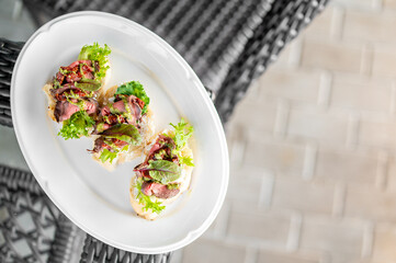 Gourmet roast beef canapés with lettuce and herbs on white plate, served on outdoor woven surface. Elegant appetizer presentation, suitable for catering or food photography