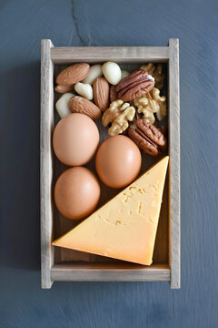 A rustic wooden crate filled with high-protein foods, including brown eggs, various nuts, and cubed cheese on a dark background