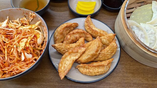 Person uses chopsticks to pick a golden fried dumpling (Gun-mandu) from a table filled with an authentic Korean feast of spicy noodles and steamed mandu in Suwon - top-down perspective