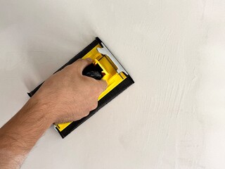 A builder smooths a wall after plastering using a sanding tool. The image depicts construction work and interior finishing in home improvement.