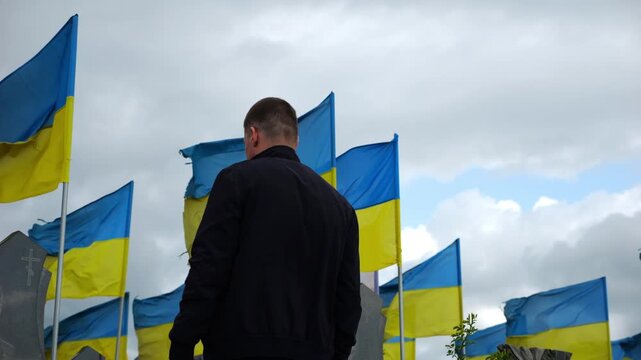 Mourning man standing among countless graves of ukrainian soldiers with national banners pay tribute to fallen family member in Kharkiv. Scene reflect personal sorrow loss caused by russian aggression
