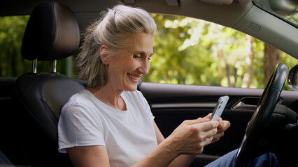 Naklejka premium Cheerful Caucasian mature woman elderly senior female retired pensioner car automobile driver inside auto using mobile phone playing game clench fist celebrating win victory betting success offer deal