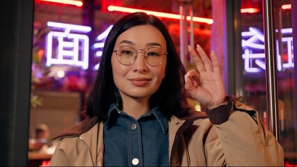Smiling Japanese business woman female Chinese businesswoman girl looking camera showing ok sign alright yes gesture outside city street neon. The inscription noodle in Chinese. Translation: 'noodle'.