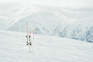 Pair of white skis with poles planted in snow on mountain slope with alpine peaks in background,...