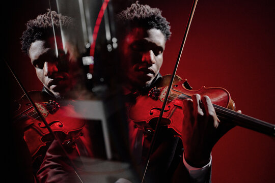 Young adult Black man playing violin with intense focus, creative mirror reflection effect showing multiple angles of musician and instrument against solid red background