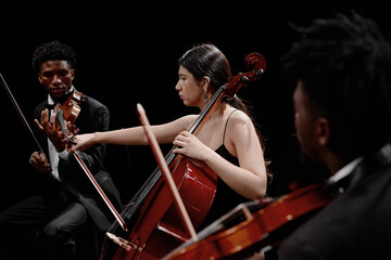 Young adult Black men playing violins with young adult Hispanic woman playing cello on stage, musicians performing classical music in ensemble
