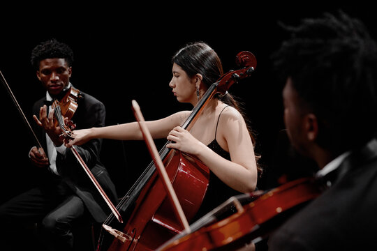 Young adult Black men playing violins with young adult Hispanic woman playing cello on stage, musicians performing classical music in ensemble