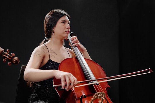 Young adult Hispanic woman playing cello on stage, focused expression, long dark hair, visible tattoo on upper arm, performing classical music in professional concert setting