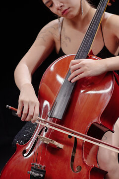 Young adult woman playing cello, focusing on hand positioning and bow technique, visible tattoos on arm, performing on stage, close up of instrument and musician