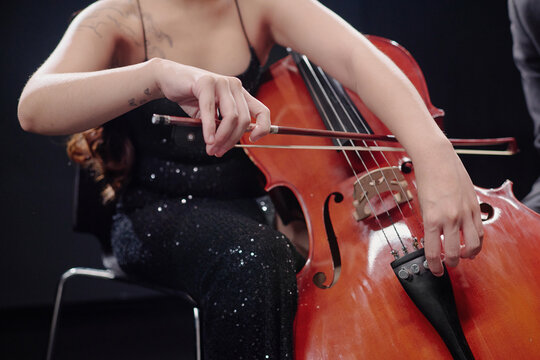 Young adult woman playing cello on stage, focusing on hand positioning and bow technique, visible tattoos on arm, wearing formal attire, seated during performance