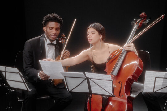 Young adult Black man playing violin and young adult Hispanic woman playing cello reading sheet music together during classical music rehearsal, both seated on stage with instruments