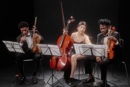 Multiethnic group of young adult musicians performing classical music on stage, Hispanic woman playing cello while two Black men playing violins, all reading sheet music together