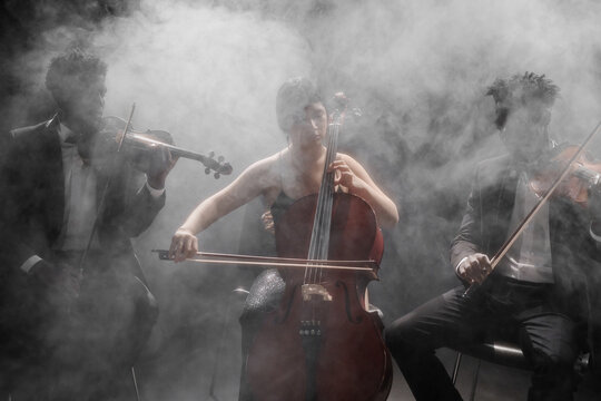 Multiethnic group of young adult musicians performing on stage, Black men playing violins and Hispanic woman playing cello, all focused on instruments with dramatic smoke background - Powered by Adobe