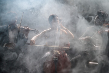 Three young adult musicians performing on stage, two Black men playing violins and one Hispanic woman playing cello, surrounded by dramatic smoke effect during live concert