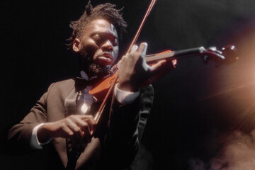 Black young adult man playing violin on stage, wearing formal suit, focusing intently on performance, dramatic lighting highlighting facial expression and instrument