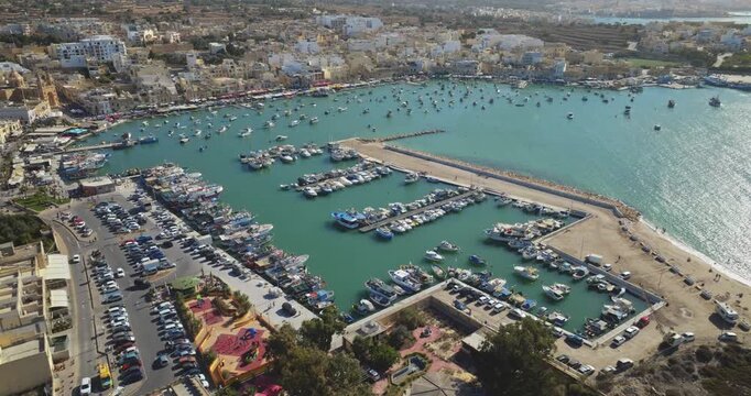 Malta: Aerial view Marsaxlokk Harbour marina bay in Malta, featuring many colorful traditional Luzzu boats moored at the docks with the town behind. Drone flight footage panorama