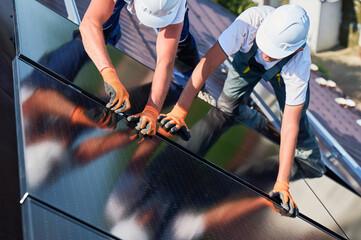 Workers building solar panel system on rooftop of house. Two men installers in helmets installing photovoltaic solar module outdoors. Alternative, green and renewable energy generation concept.