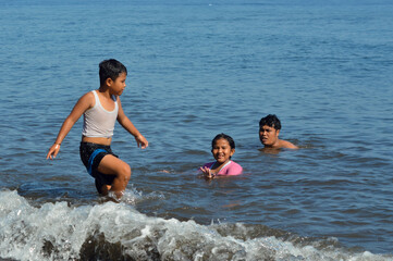 A young boy walks excitedly toward the sandy shore while his father and sister enjoy bathing together in the shallow sea on a bright tropical day.
