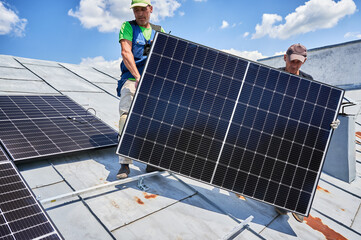Workers building solar panel system on metal rooftop of house. Two men installers carrying photovoltaic solar module outdoors. Alternative, green and renewable energy generation concept.