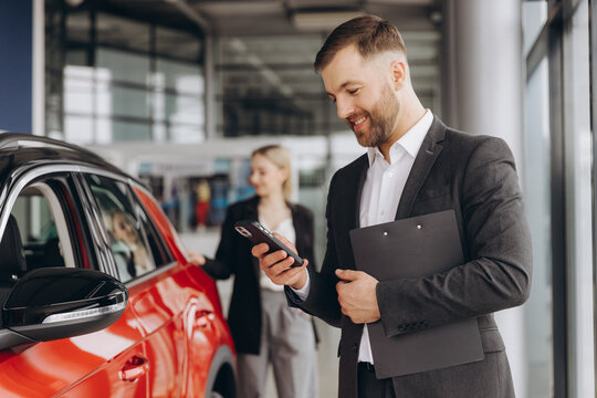 Smiling salesman using smartphone while having business call at car showroom. Car dealer holding clipboard in automobile showroom. Professional confident sales person working in modern car dealership.