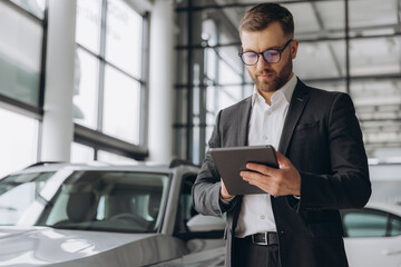 Bearded friendly car seller in glasses and suit standing in car salon and holding tablet