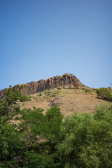 Scenic view of a rocky mountain ridge with unique basalt formations, surrounded by dry grassy slopes and lush green trees. A natural landscape under a bright blue sky, geology, travel and adventure