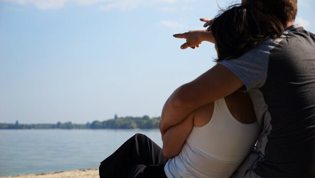 Happy pair hugs during sitting on wooden log on beach. Romantic couple admiring sea landscape at sunny day. Lovers spending time together at resort. Concept of summer vacation or holiday. Slow mo - Powered by Adobe