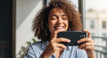 Joyful african american woman capturing moments with her smartphone indoors in natural light smiling pretty with curly hair