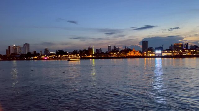 Scenic view of Phnom Penh riverside skyline at twilight, with illuminated buildings, temple architecture, boats, and colorful reflections shimmering on the Tonle Sap River