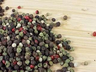 A mixture of various peppercorns scattered across a light wooden surface. The black, white, green, and red peppercorns are visible, creating a contrasting and textured image of the spice.