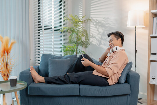 A young man relaxes at home, smiling while using his smartphone. He enjoys leisure time with headphones around his neck, sitting comfortably and feeling casual in a modern living room.