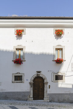 View of a bright white building facade adorned with vibrant red flowers in window boxes, framed by an arched doorway, Pescocostanzo, Abruzzo, Italy.
