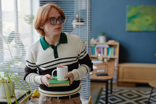 Young adult Caucasian man with red hair and glasses holding coffee mug and book standing in modern office space, looking away from camera with thoughtful expression