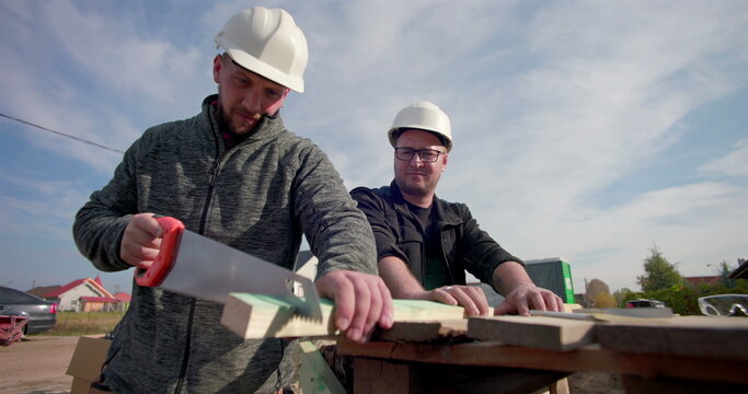 Construction worker cutting wood plank with handsaw, colleague observing, teamwork on construction site, house framework visible, focused on precision and craftsmanship