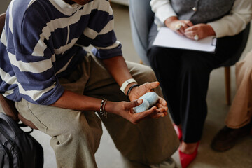 Obraz premium Black man sitting in group therapy session holding stress balls in hands while counselor with clipboard and another participant listening during discussion
