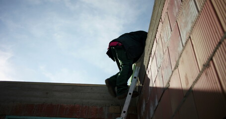 Construction workers descending ladder carefully on unfinished building site, maintaining safety protocols, brick walls and window frame visible in background