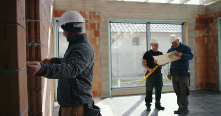 Construction worker using measuring tape on brick wall, background shows team discussing blueprints...